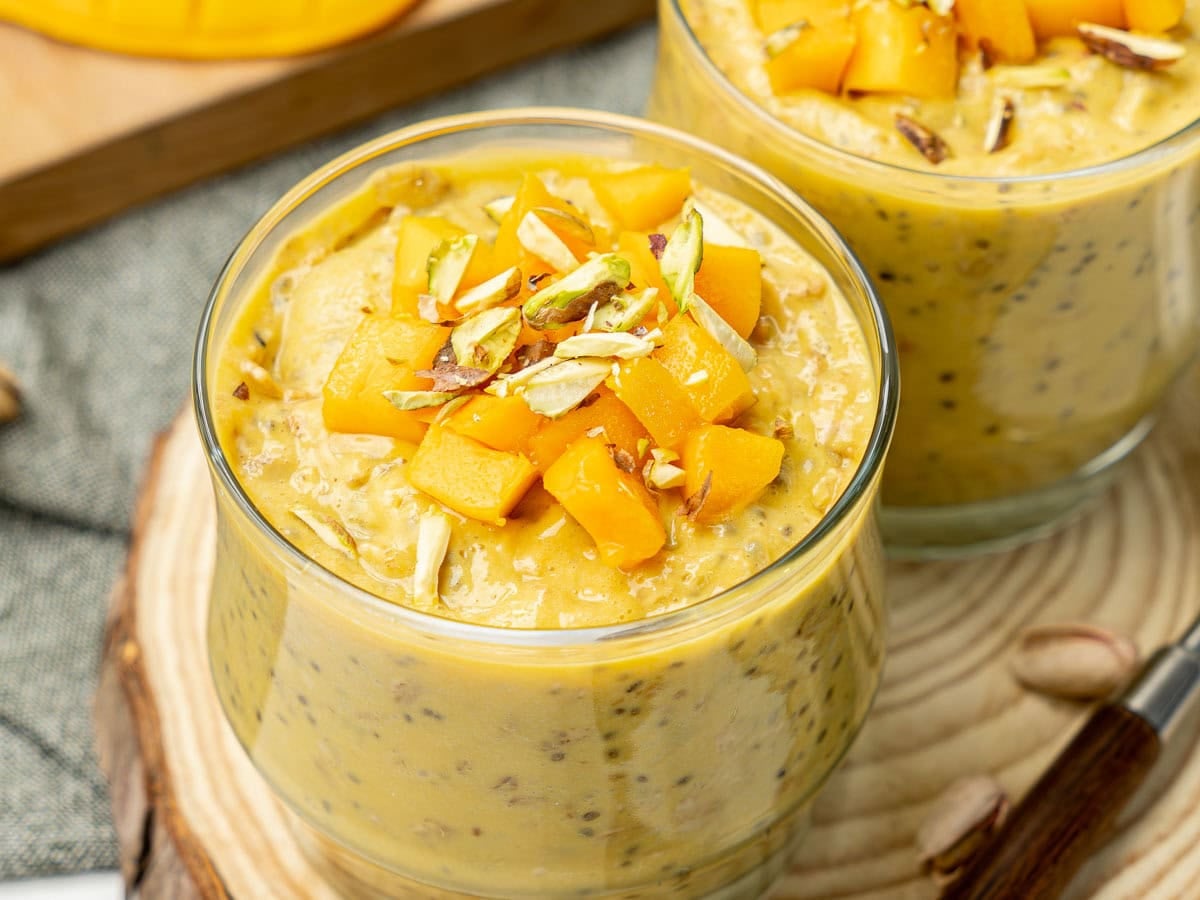Creamy mango overnight oats served in two cups, topped with fresh mango cubes and slivered pistachios.