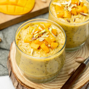Mango overnight oats served in two cups for breakfast, topped with fresh mango cubes and slivered pistachios.