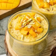 Mango overnight oats with greek yogurt served in two cups, topped with fresh mango cubes and slivered pistachios.