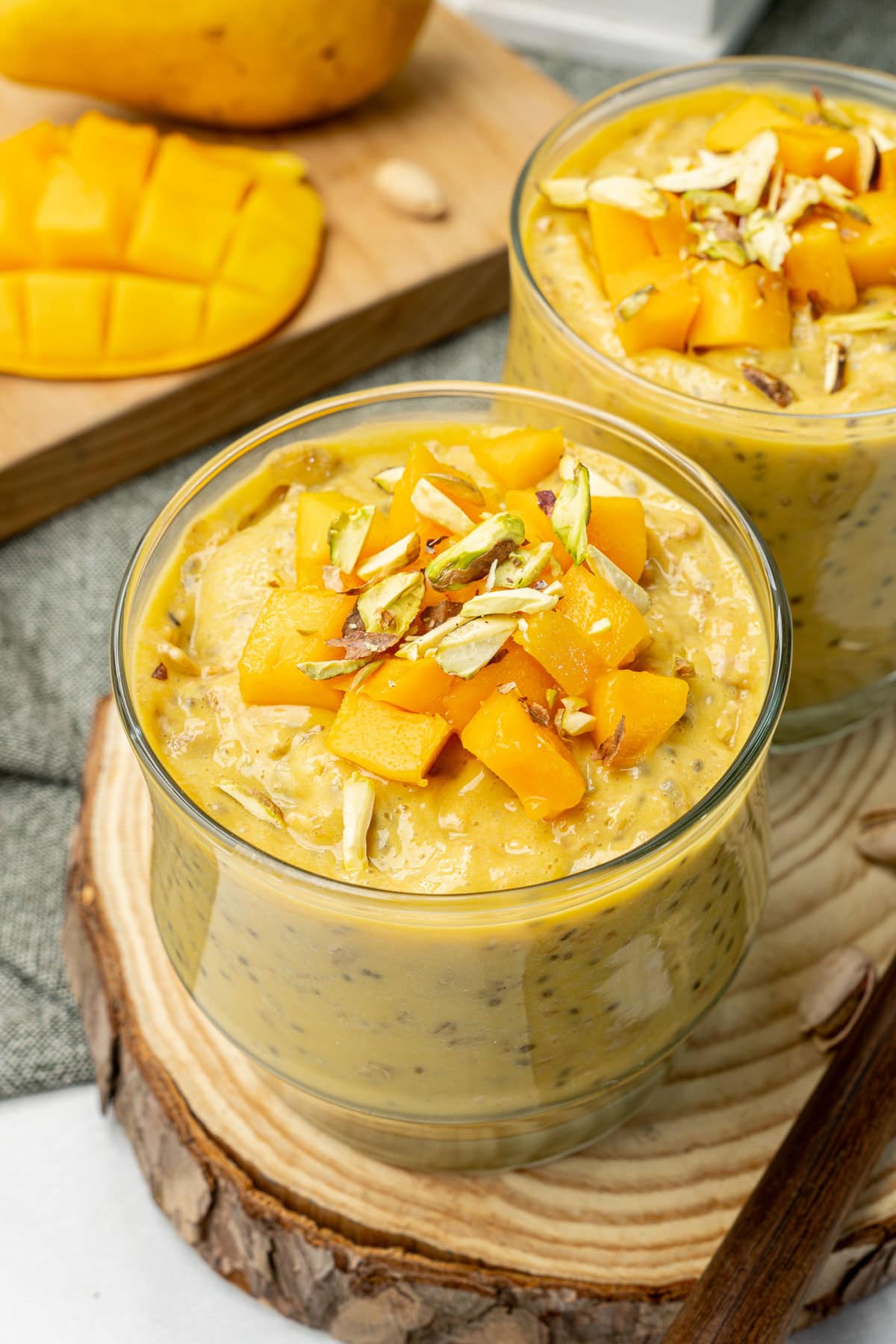 Mango overnight oats with greek yogurt served in two cups, topped with fresh mango cubes and slivered pistachios.